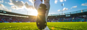 A low-angle close-up of a football player's cleat on a pitch, with sunlight flaring behind it and a stadium in the background.